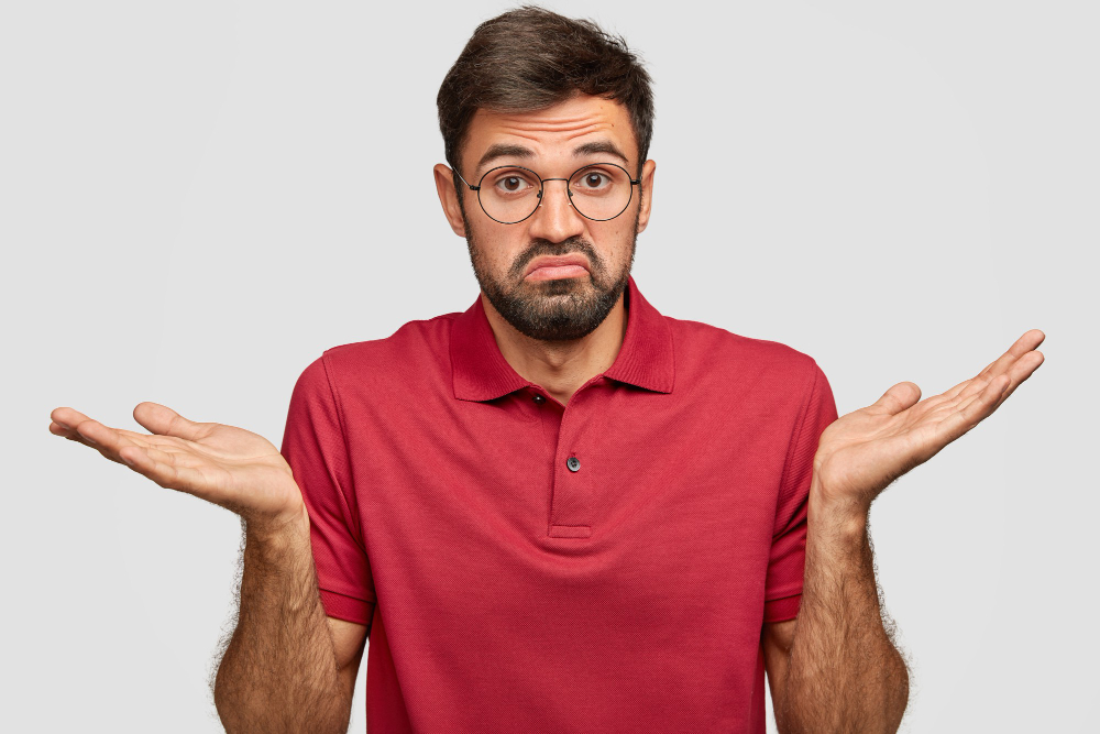 Indoor shot of good looking puzzled confused young male with dark beard, feels hesitant, raises palms with bewilderment, says: Maybe yes or no, wears casual red tshirt, isolated over white wall